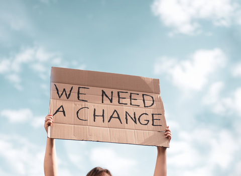A person holds up a sign made out of cardboard which reads: "WE NEED A CHANGE."