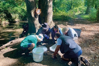A group of people surrounding survey equipment beside a small brook