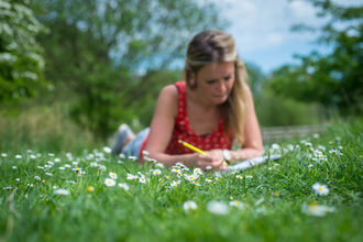 a lady laying on grass and daisies with a notebook and pencil