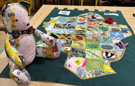 Photo of Lemmy the Leamington Patchwork bear with a collection of fabrics used and the patchwork artwork itself with different wildlife in the area
