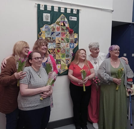 Group of ladies stood in front of the patchwork map they sewed with bunches of flowers