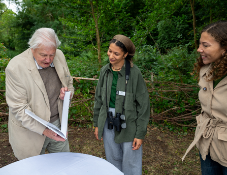 Sir David Attenborough stood with 2 people as he holds a book open