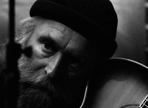 Close up black and white photo of a man with a hat, beard and guitar.