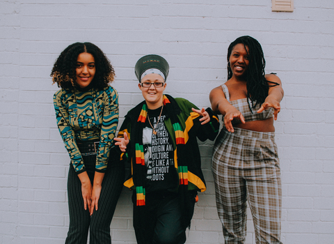 Group of 3 women dressed colourfully against a white brick wall